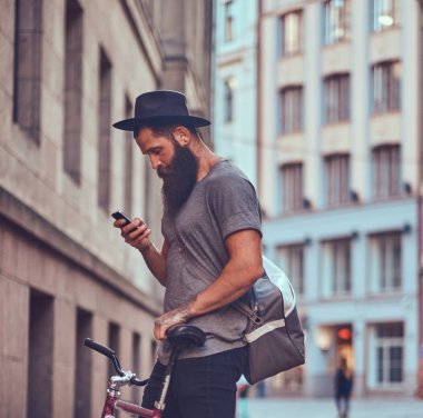 Bir şık sakal ve kollarını bir çanta ile rahat kıyafetler giymiş dövme ile bir yakışıklı hipster seyahat standları kaldırımda Bisiklete binmek sonra dinlenme.