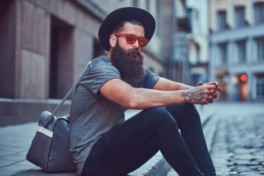 Bir yakışıklı hipster seyahat bir şık sakal ve kollarını bir çanta ile rahat kıyafetler giymiş dövmesini oturur kaldırımda Bisiklete binmek sonra dinlenme.