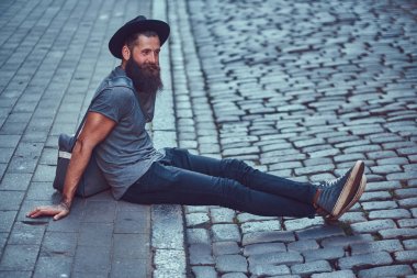Bir yakışıklı hipster seyahat bir şık sakal ve kollarını bir çanta ile rahat kıyafetler giymiş dövmesini oturur kaldırımda Bisiklete binmek sonra dinlenme.
