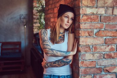 Bir t-shirt ve şapka bir tuğla duvara poz tattoed seksi sarışın.