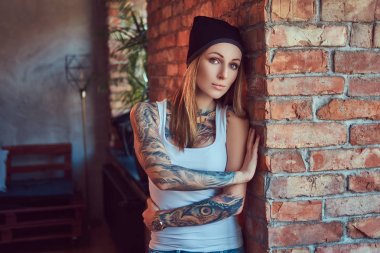 Bir t-shirt ve şapka bir tuğla duvara poz tattoed seksi sarışın.