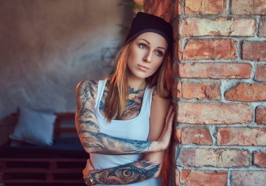 Bir t-shirt ve şapka bir tuğla duvara poz tattoed seksi sarışın.