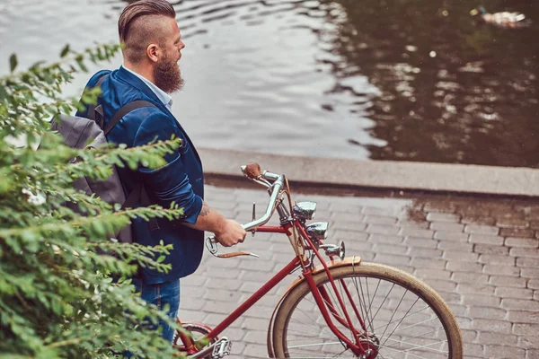 Homem barbudo com um corte de cabelo elegante vestido com roupas casuais com uma mochila, de pé com uma bicicleta retro perto do rio em um parque da cidade . — Fotografia de Stock