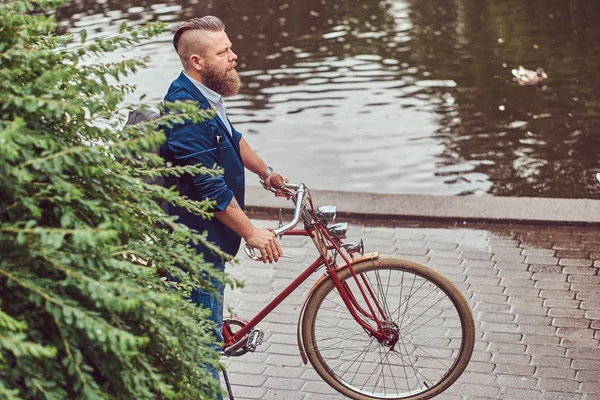 Bärtiger Mann mit stylischem Haarschnitt in lässiger Kleidung mit Rucksack, der mit einem Retro-Fahrrad in Flussnähe in einem Stadtpark steht. — Stockfoto