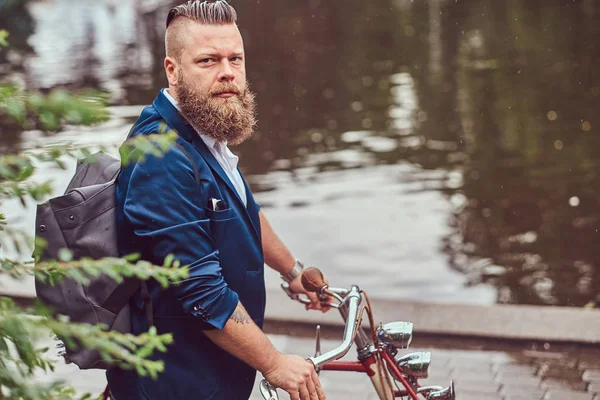 Бородатий чоловік зі стильною зачіскою, одягнений у повсякденний одяг з рюкзаком, стоїть з ретро велосипедом біля річки в міському парку . — стокове фото