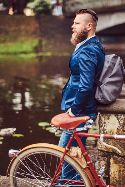 Varón barbudo con un corte de pelo elegante vestido con ropa casual con una mochila, de pie con una bicicleta retro cerca del río en un parque de la ciudad . — Foto de Stock