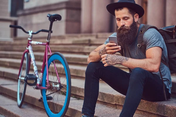 A handsome hipster traveler with a stylish beard and tattoo on his arms dressed in casual clothes, sitting on the steps, using the phone, resting after a bike ride. — Stock Photo, Image