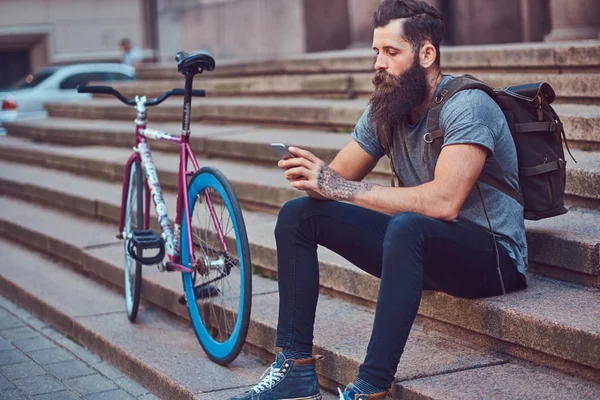Красивый хипстер-путешественник со стильной бородой и татуировкой на руках, одетый в повседневную одежду, сидящий на ступеньках, пользующийся телефоном, отдыхающий после езды на велосипеде . — стоковое фото