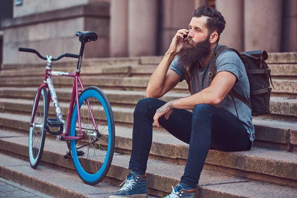 Ένας όμορφος hipster ταξιδιώτης με στιλάτα γένια και τατουάζ στα χέρια του, ντυμένοι με casual ρούχα, καθισμένος στα σκαλιά, χρησιμοποιώντας το τηλέφωνο, ανάπαυση μετά από μια βόλτα με ποδήλατο. — Φωτογραφία Αρχείου