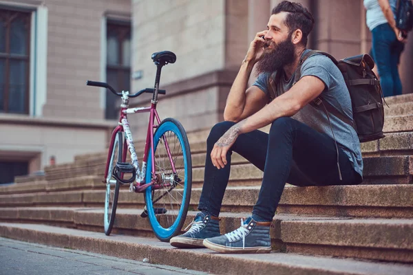 Ένας όμορφος hipster ταξιδιώτης με στιλάτα γένια και τατουάζ στα χέρια του, ντυμένοι με casual ρούχα, καθισμένος στα σκαλιά, χρησιμοποιώντας το τηλέφωνο, ανάπαυση μετά από μια βόλτα με ποδήλατο. — Φωτογραφία Αρχείου