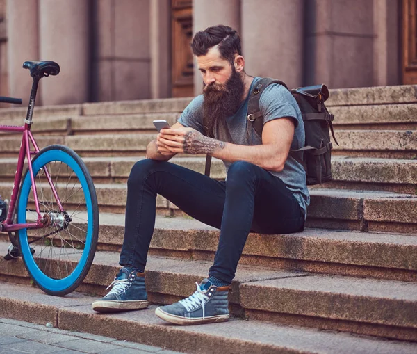 A handsome hipster traveler with a stylish beard and tattoo on his arms dressed in casual clothes, sitting on the steps, using the phone, resting after a bike ride. — Stock Photo, Image