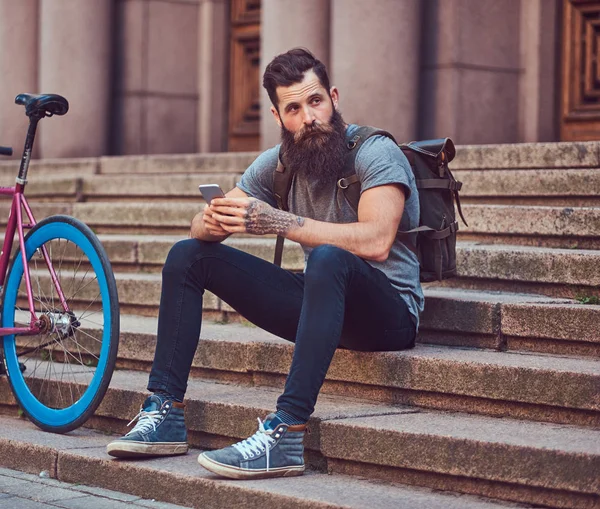 A handsome hipster traveler with a stylish beard and tattoo on his arms dressed in casual clothes, sitting on the steps, using the phone, resting after a bike ride. — Stock Photo, Image