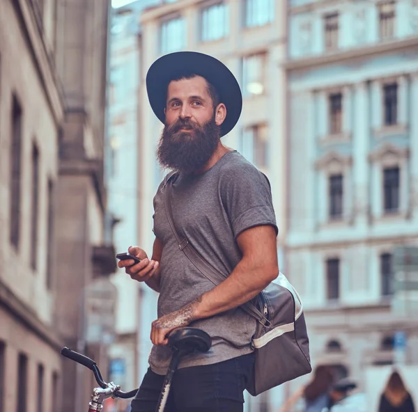 Красивый хипстер-путешественник со стильной бородой и татуировкой на руках, одетый в повседневную одежду с сумкой, стоит на тротуаре, отдыхает после велосипедной прогулки . — стоковое фото