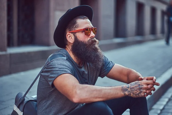 Un guapo viajero hipster con una elegante barba y tatuaje en sus brazos vestido con ropa casual con una bolsa, se sienta en la acera, descansando después de un paseo en bicicleta . — Foto de Stock