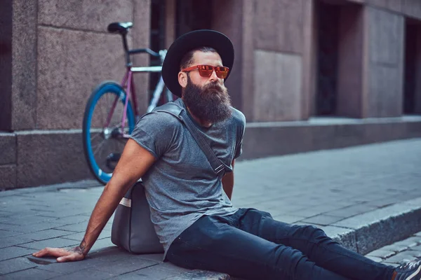 A handsome hipster traveler with a stylish beard and tattoo on his arms dressed in casual clothes with a bag, sits on the sidewalk, resting after a bike ride. — Stock Photo, Image