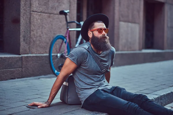 Ένας όμορφος hipster ταξιδιώτης με στιλάτα γένια και τατουάζ στα χέρια του, ντυμένοι με casual ρούχα με μια τσάντα, κάθεται στο πεζοδρόμιο, ανάπαυση μετά από μια βόλτα με ποδήλατο. — Φωτογραφία Αρχείου