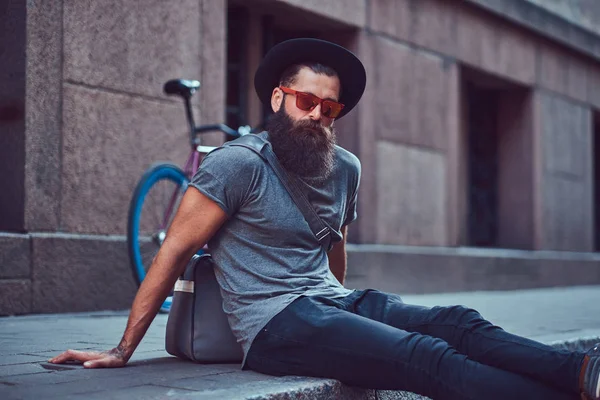 Ένας όμορφος hipster ταξιδιώτης με στιλάτα γένια και τατουάζ στα χέρια του, ντυμένοι με casual ρούχα με μια τσάντα, κάθεται στο πεζοδρόμιο, ανάπαυση μετά από μια βόλτα με ποδήλατο. — Φωτογραφία Αρχείου