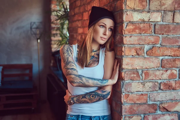 Uma loira sexy tatuada em uma camiseta e um chapéu posando contra uma parede de tijolo . — Fotografia de Stock