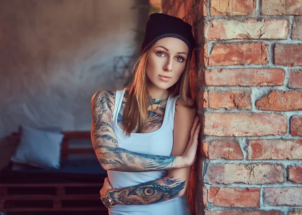 Bir t-shirt ve şapka bir tuğla duvara poz tattoed seksi sarışın.