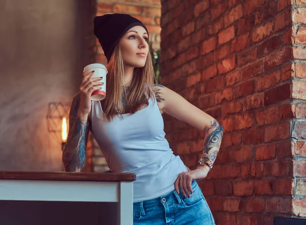 Tattoed blond sexy tričko a čepici s šálkem kávy v místnosti s interiér podkroví. — Stock fotografie