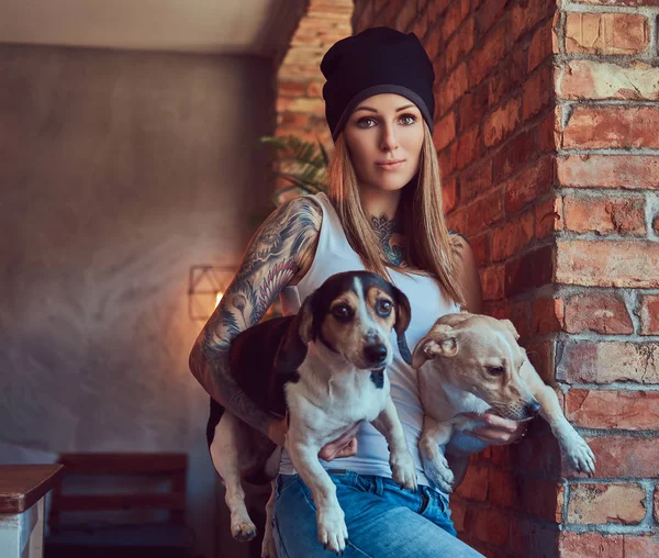 Una mujer rubia tatuada con estilo en camiseta y jeans sostiene a dos perros lindos . —  Fotos de Stock