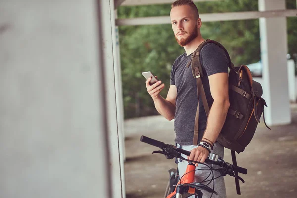 Красивий рудий чоловік зі стильною зачіскою і бородою, одягнений у спортивний одяг з велосипедом і рюкзаком, використовуючи смартфон, що стоїть зовні . — стокове фото