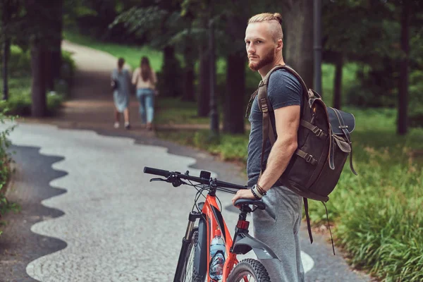 A handsome redhead male with a stylish haircut and beard dressed in casual clothes walks in the park with a bicycle and backpack.