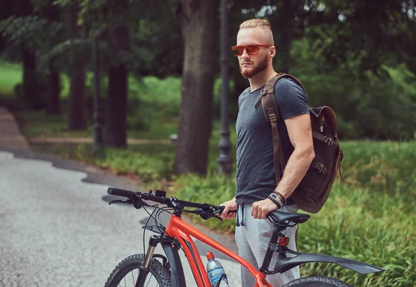 Un hombre pelirrojo guapo con un corte de pelo elegante y barba vestida con ropa deportiva y gafas de sol camina por el parque con una bicicleta y una mochila. — Foto de Stock