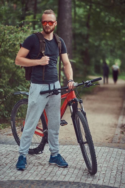 Красивый рыжий мужчина со стильной стрижкой и бородой, одетый в спортивную одежду и солнечные очки, прогуливается по парку с велосипедом и рюкзаком. — стоковое фото