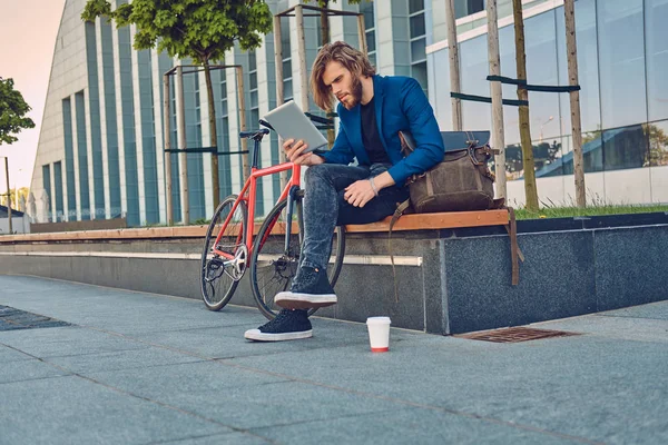 Un guapo hombre barbudo de moda con el pelo largo sentado en un banco con una bicicleta, usando una tableta . — Foto de Stock