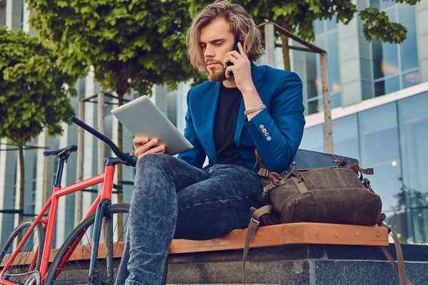 A handsome fashionable bearded male with long hair sitting on a bench with a bicycle, working with a tablet computer and speak on the phone. — Stock Photo, Image
