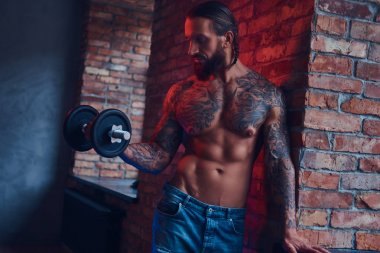 Bir çatı katı iç ile bir odada bir tuğla duvara duran bir dumbbell ile egzersiz yaparak bir yakışıklı tattoed gömleksiz olmayan erkek bir şık saç ve sakal,.