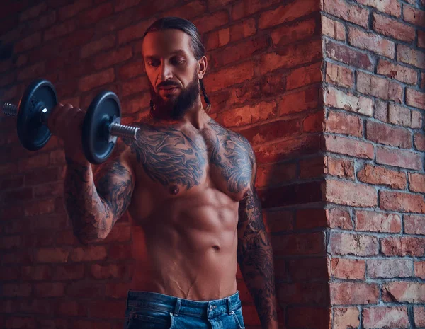 Um belo homem sem camisa tatuado com um corte de cabelo elegante e barba, fazendo exercício com um halteres, de pé contra uma parede de tijolos em uma sala com um interior loft . — Fotografia de Stock
