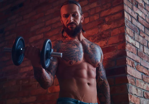 Um belo homem sem camisa tatuado com um corte de cabelo elegante e barba, fazendo exercício com um halteres, de pé contra uma parede de tijolos em uma sala com um interior loft . — Fotografia de Stock