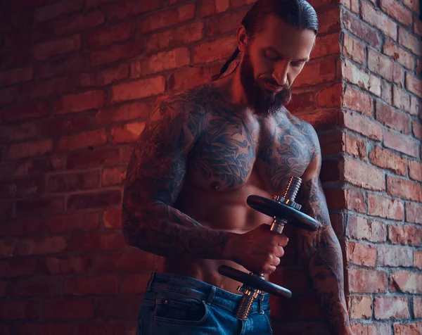 Ένα όμορφος tattoed shirtless αρσενικό με ένα μοντέρνο κούρεμα και γενειάδα, κάνει άσκηση με έναν αλτήρα, στέκεται ενάντια σε έναν τοίχο από τούβλα σε ένα δωμάτιο με ένα εσωτερικό πατάρι. — Φωτογραφία Αρχείου