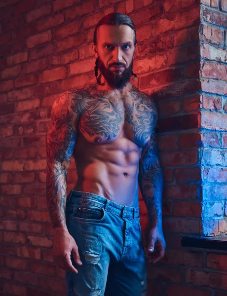 Een knappe tattoed shirtless man met een stijlvol kapsel en baard, staande tegen een bakstenen muur in een kamer met een loft interieur. — Stockfoto