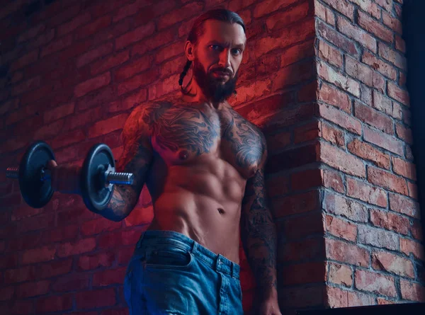 Un guapo hombre sin camisa tatuado con un corte de pelo elegante y barba, haciendo ejercicio con una mancuerna, de pie contra una pared de ladrillo en una habitación con un interior loft . —  Fotos de Stock