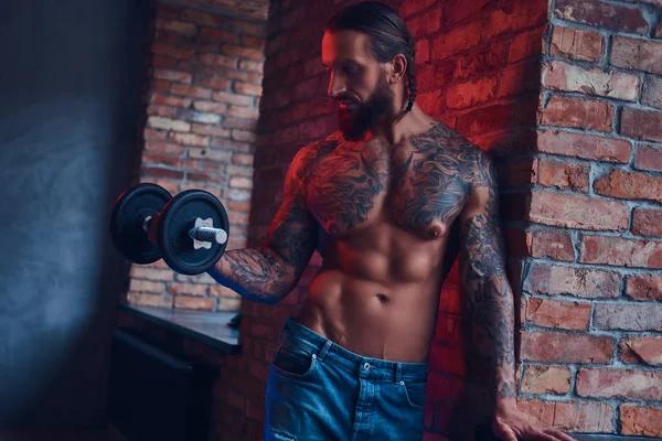 Un guapo hombre sin camisa tatuado con un corte de pelo elegante y barba, haciendo ejercicio con una mancuerna, de pie contra una pared de ladrillo en una habitación con un interior loft . — Foto de Stock