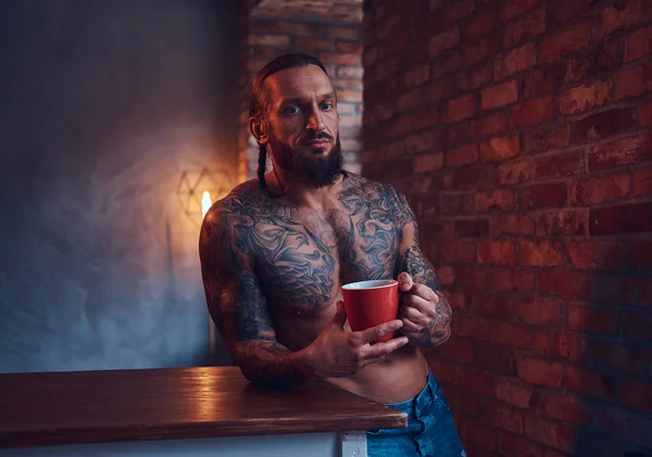Un hombre guapo barbudo tatuado con un corte de pelo elegante y cuerpo musculoso, bebe café, apoyado en una mesa . — Foto de Stock