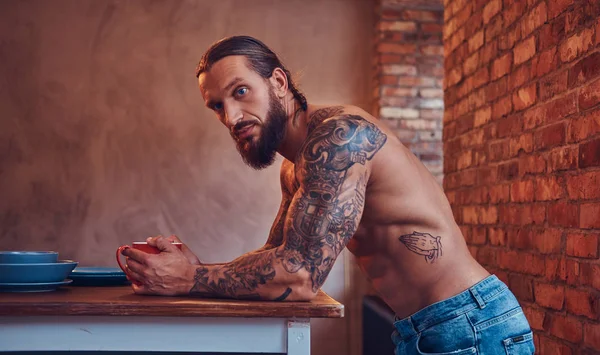 Um belo homem de barba tatuado com um corte de cabelo elegante e corpo muscular, bebe café, encostado em uma mesa . — Fotografia de Stock