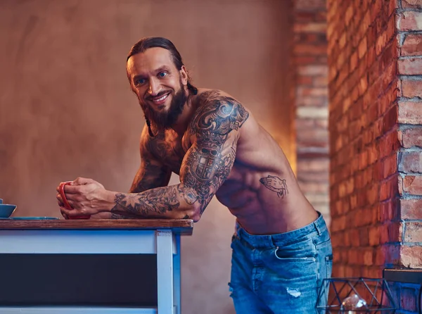 Un beau mâle tatoué barbu avec une coupe de cheveux élégante et un corps musclé, boit du café, appuyé sur une table . — Photo