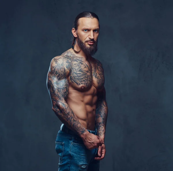 Close-up portret van een gespierde naakte bebaarde tattoed man met een stijlvol kapsel, geïsoleerd op een donkere achtergrond. — Stockfoto