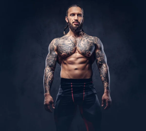 Retrato de un macho musculoso con barba desnuda tatuado con un corte de pelo elegante en pantalones deportivos, aislado sobre un fondo oscuro . —  Fotos de Stock