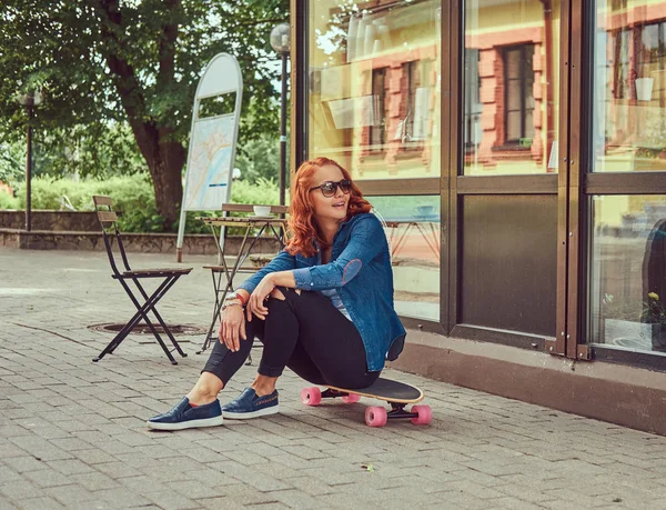 Een jonge roodharige meisje met een skateboard, zitten in de buurt van een coffeeshop. — Stockfoto