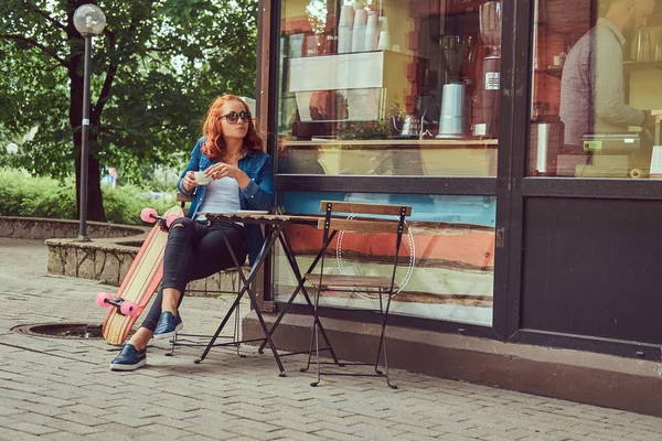 Ein junges rothaariges Mädchen trinkt einen Kaffee, sitzt in der Nähe eines Coffeeshops und entspannt sich nach dem Skateboardfahren. — Stockfoto