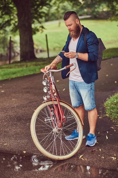 Путешественник, одетый в повседневную одежду с рюкзаком, с помощью смартфона, отдыхающий в городском парке после езды на ретро-велосипеде . — стоковое фото