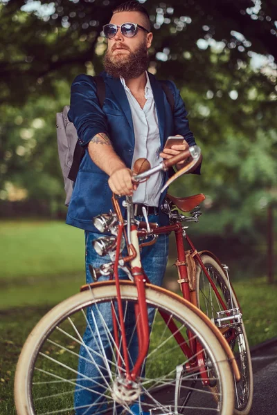 Um viajante vestido com roupas casuais e óculos de sol com uma mochila, usando um smartphone, relaxando em um parque da cidade depois de andar em uma bicicleta retro . — Fotografia de Stock