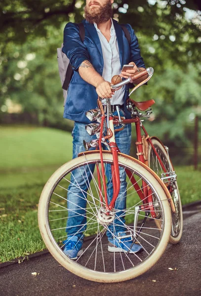 Een reiziger gekleed in casual kleding en zonnebrillen met een rugzak, met behulp van een smartphone, ontspannen in een stadspark na het rijden op een retro fiets. — Stockfoto