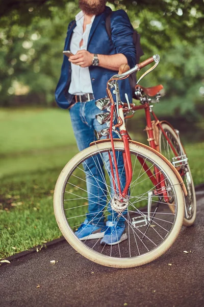 Ein Reisender in lässiger Kleidung und Sonnenbrille mit einem Rucksack, der ein Smartphone benutzt und sich nach der Fahrt mit einem Retro-Fahrrad in einem Stadtpark entspannt. — Stockfoto