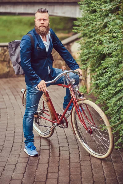 Бородатий чоловік зі стильною стрижкою, одягнений у повсякденний одяг з рюкзаком, сидить на ретро-велосипеді в міському парку . — стокове фото